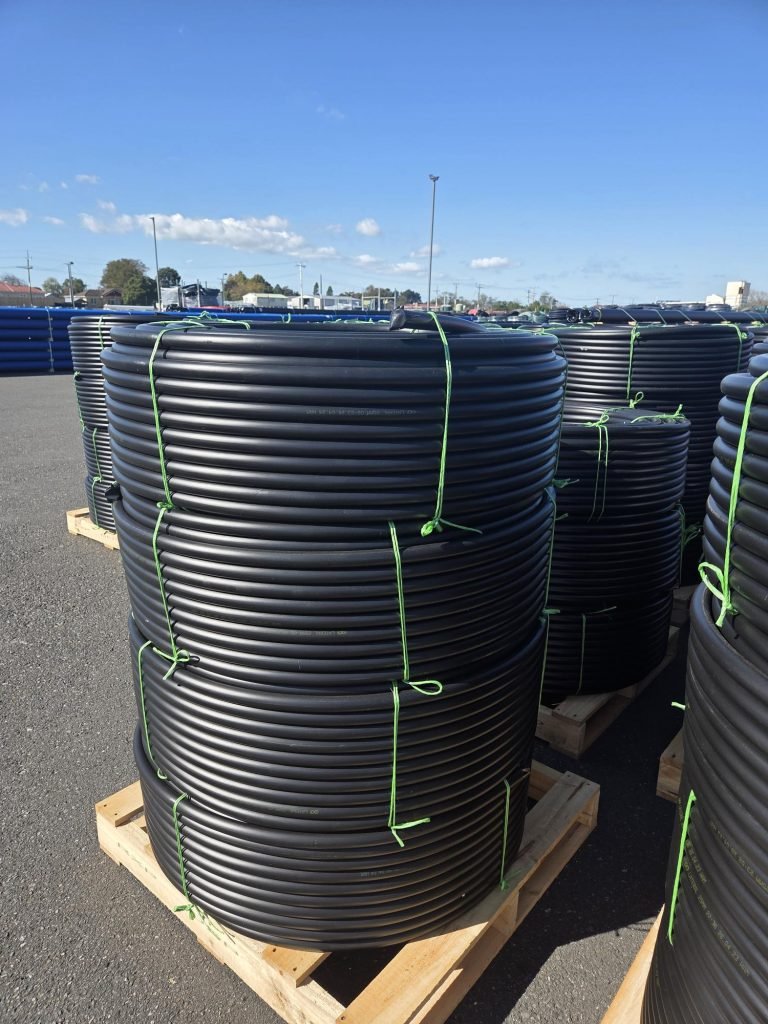 Coiled black plastic tubing stacked on wooden pallets in an outdoor storage lot with green tying straps on a sunny day.